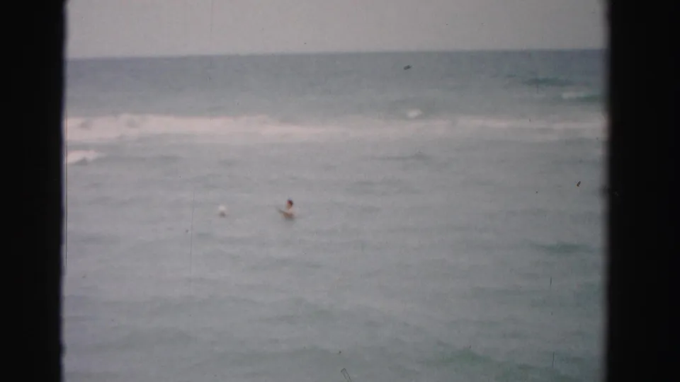1955 Florida beach home-movie frame showing distant swimmers bobbing in the ocean near low breaking waves.