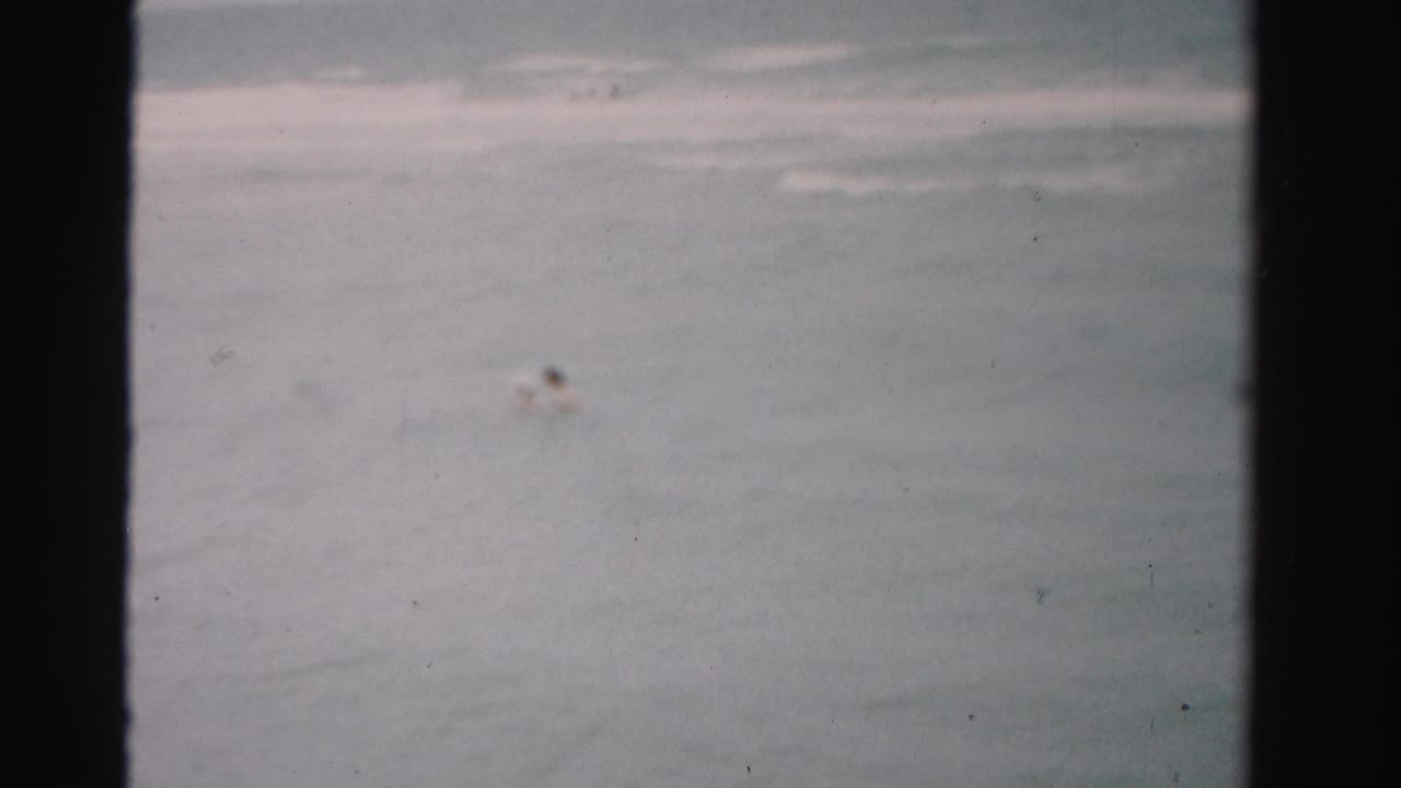 1955 Florida home-movie frame showing a distant swimmer in gray-blue water with low surf behind them.