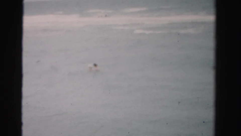 1955 Florida home-movie frame showing a distant swimmer in gray-blue water with low surf behind them.
