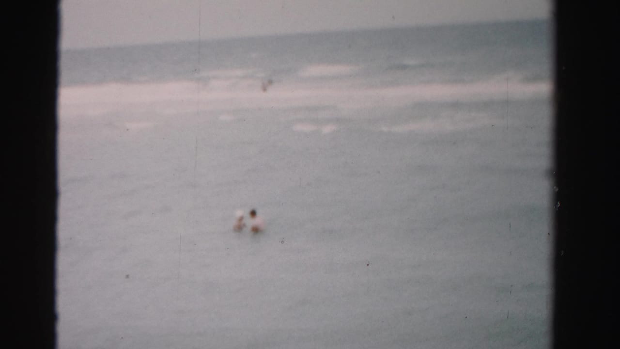 1955 Dade County 8mm frame showing a small swimmer offshore in muted gray-blue water under an overcast sky.