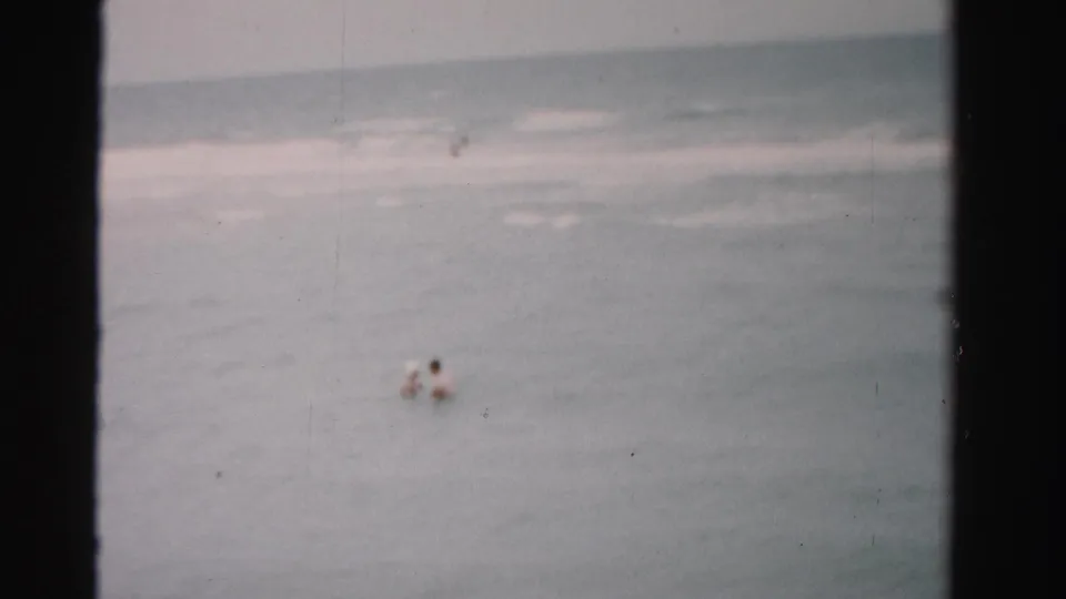 1955 Dade County 8mm frame showing a small swimmer offshore in muted gray-blue water under an overcast sky.