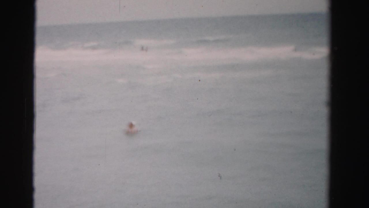 1955 Florida home-movie frame with a distant swimmer in open water and muted surf near the horizon.