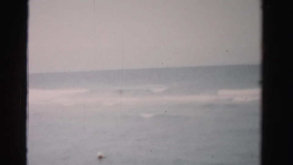 1955 Dade County home-movie frame showing pale ocean surf, low horizon, and dark 8mm gate edges.