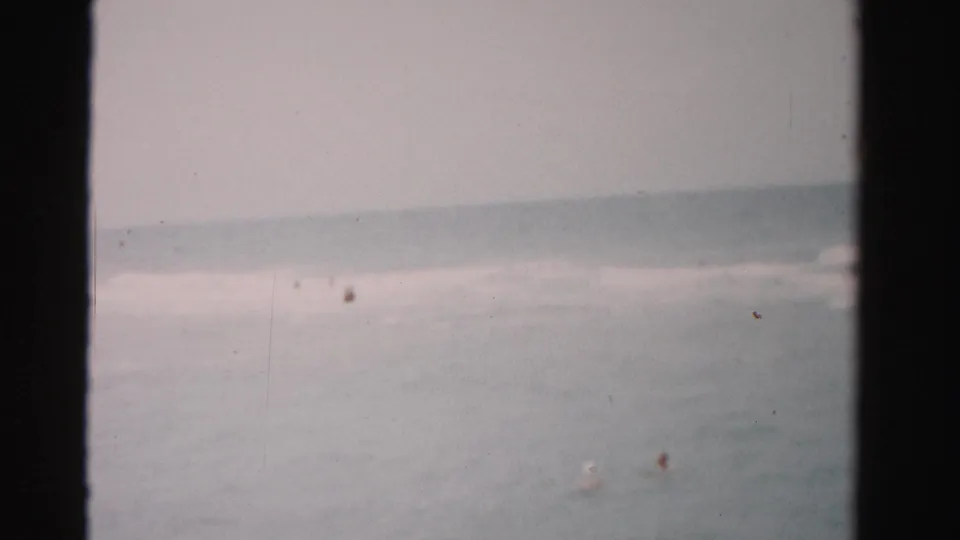1955 Dade County surf scene with a low horizon, pale water, and visible 8mm film texture.