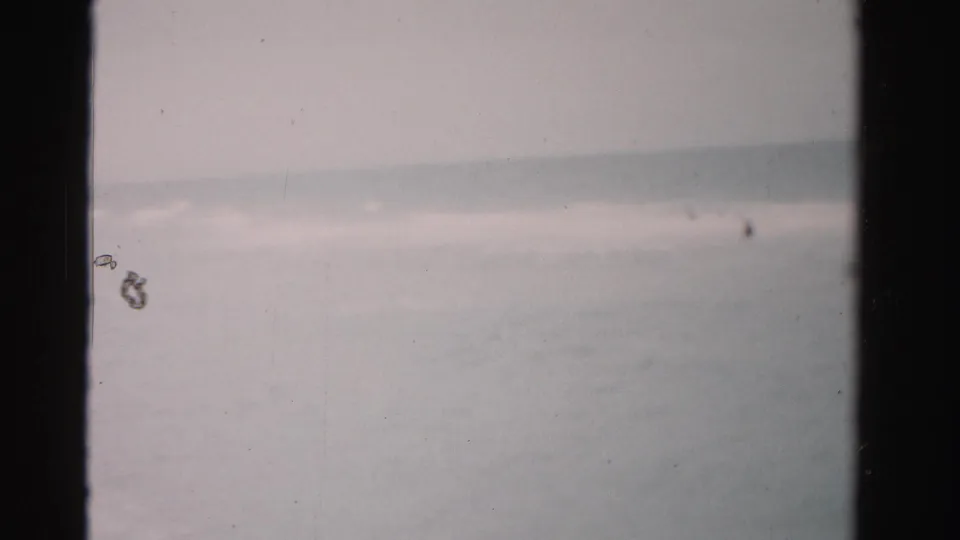 1955 Dade County, Florida home-movie frame showing muted surf, a pale horizon, and tiny distant swimmers within dark film borders.