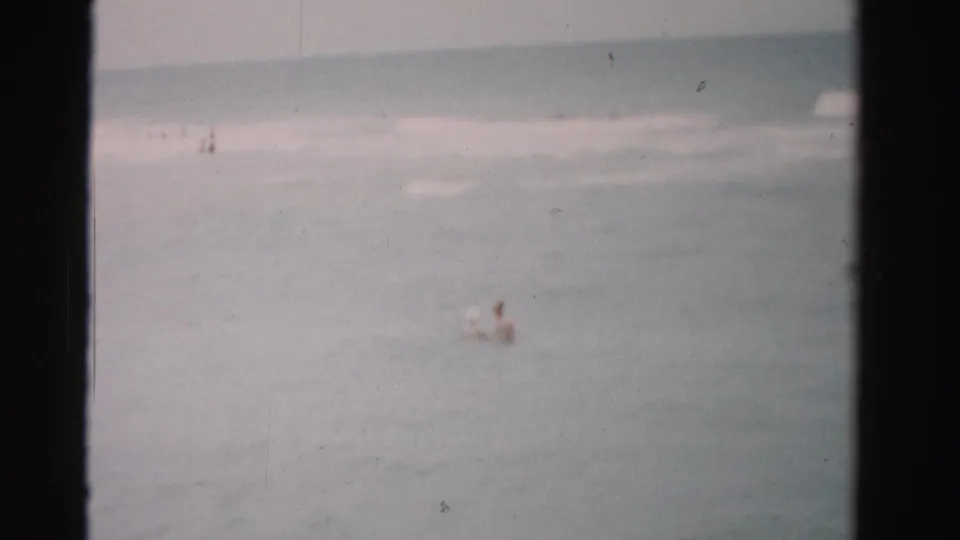 Muted 1955 Florida beach footage shows distant bathers or swimmers in soft focus beyond low surf and a hazy shoreline.