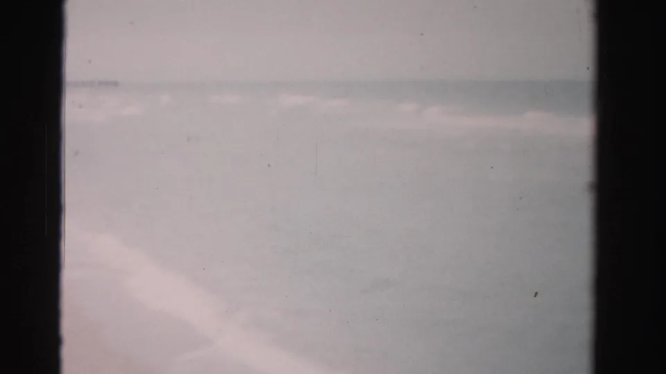 A 1955 Dade County beach view shows pale sand, gentle waves, and a soft horizon in a grainy 8mm home-movie frame.