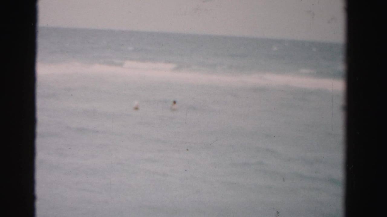 1955 Dade County 8mm frame showing distant swimmers in open ocean with low surf and a pale horizon.