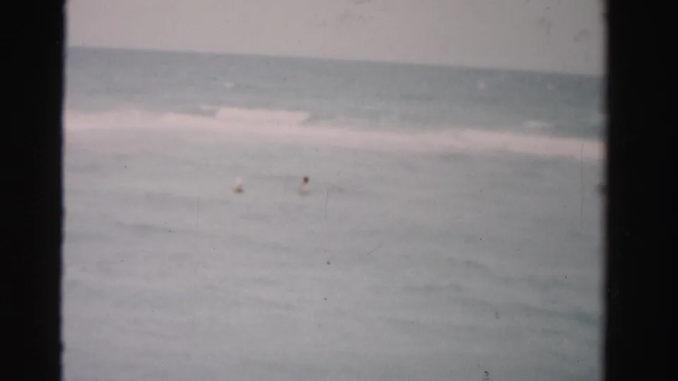 1955 Dade County 8mm frame showing distant swimmers in open ocean with low surf and a pale horizon.