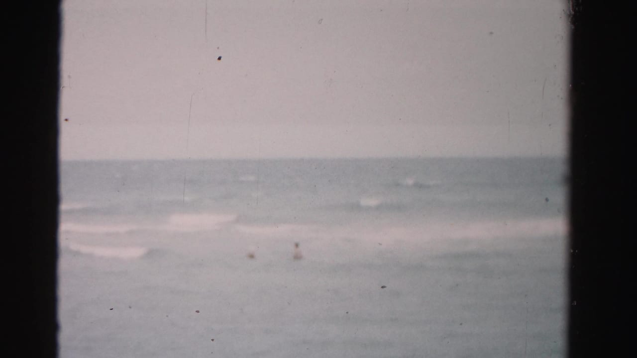 Open ocean with faint surf lines and tiny distant figures in the water.