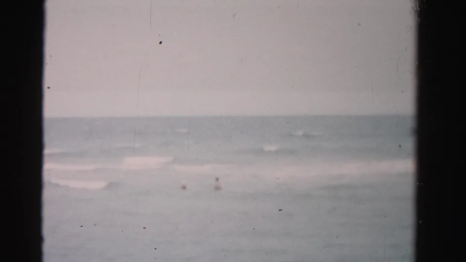 1955 Dade County beach footage frame with tiny swimmers barely visible beyond soft bands of surf.