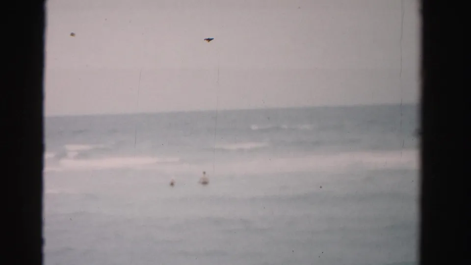 Faded 8mm frame from Dade County showing small swimmers in the surf under a muted sky.