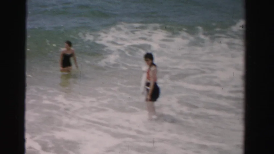 In 1955 Dade County beach footage, two women wade in shallow ocean water as white surf rolls toward shore.