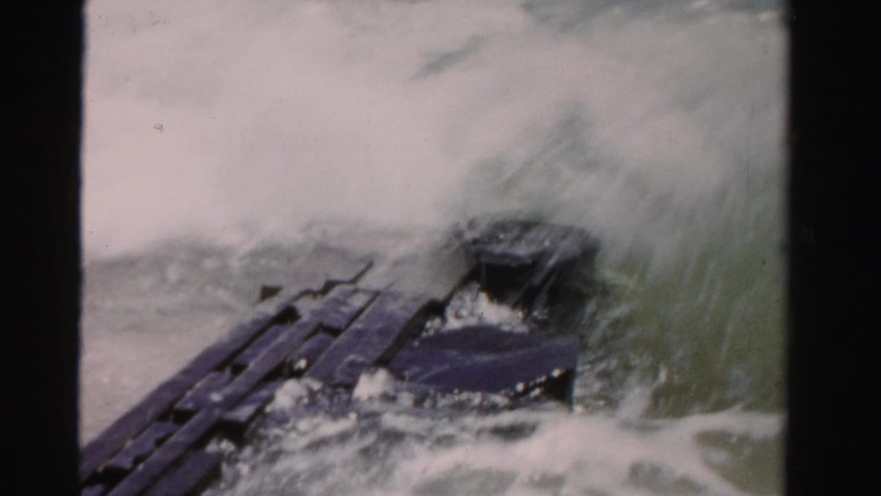 Breaking wave crashes over a dark elongated structure in the water.