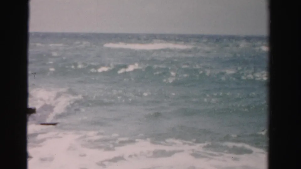 1955 Dade County, Florida 8mm frame showing rough ocean waves and white surf near a rocky jetty, viewed through a dark window-like frame.