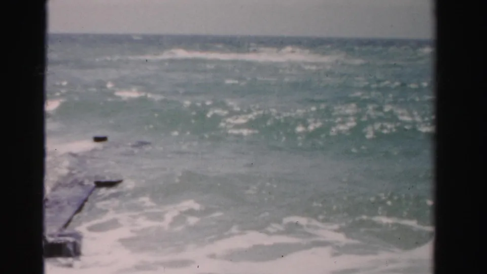 1955 Dade County coastal frame with choppy surf, a narrow rocky jetty, and dark foreground framing from a window or doorway.