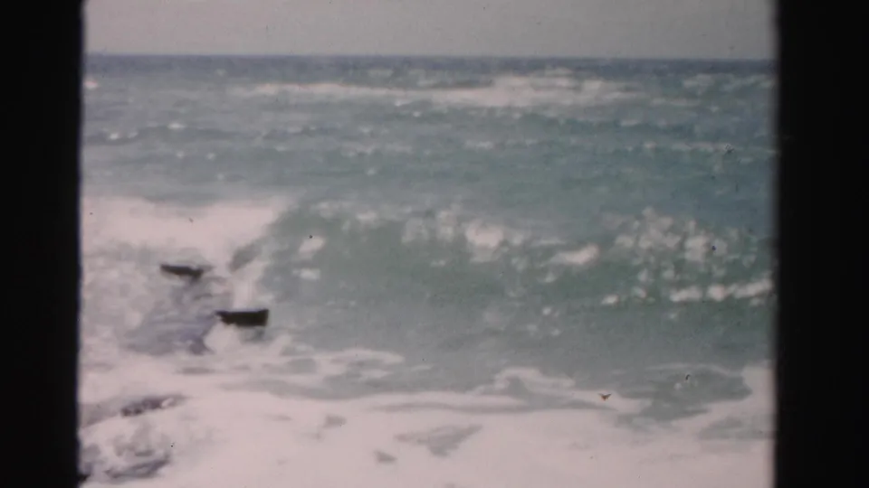 1955 Dade County 8mm frame showing foamy Atlantic surf and rocks near shore, partially obscured by dark foreground framing.