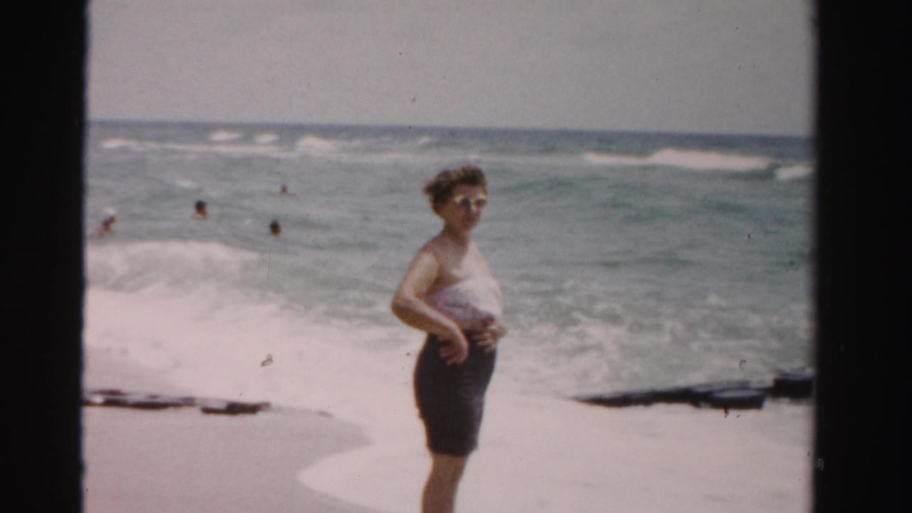 Woman in a white top and dark shorts standing on a beach with ocean waves behind her.
