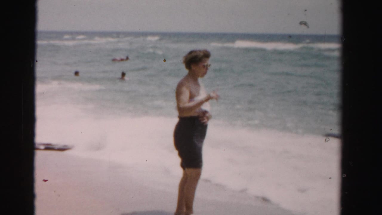 Woman in profile near foamy surf with swimmers visible in the ocean.
