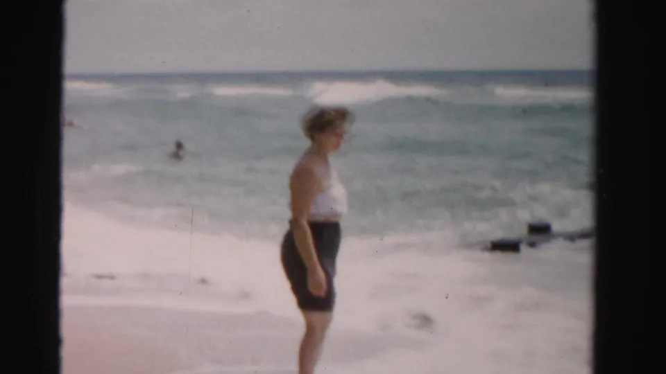 Soft 8mm frame from 1955 Dade County, Florida showing a woman at the beach edge with ocean surf and a swimmer offshore.