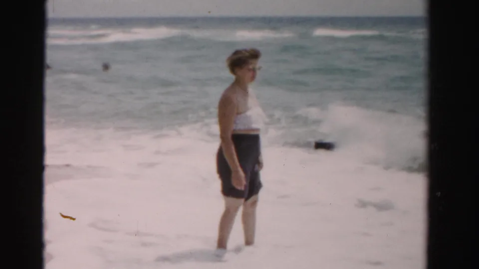 1955 Dade County, Florida 8mm still with a woman standing on wet sand as white surf breaks close behind her.