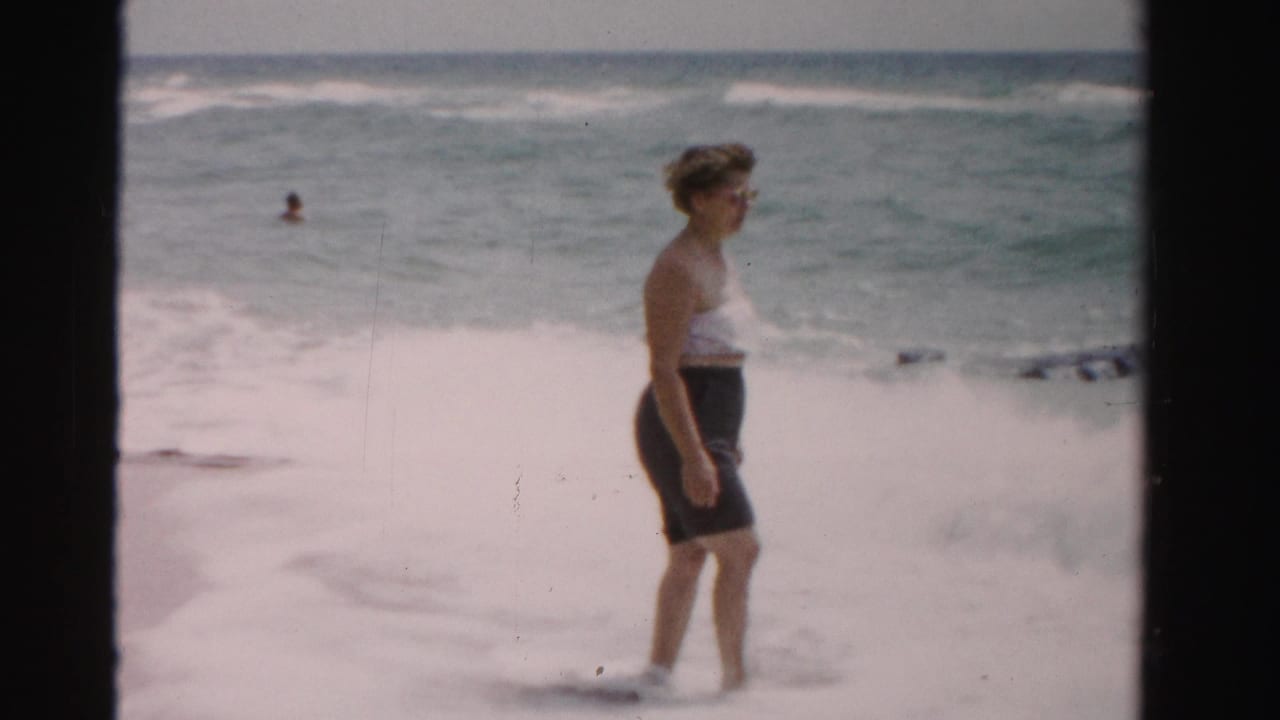 Woman in sunglasses stands in shallow surf with ocean waves behind her.