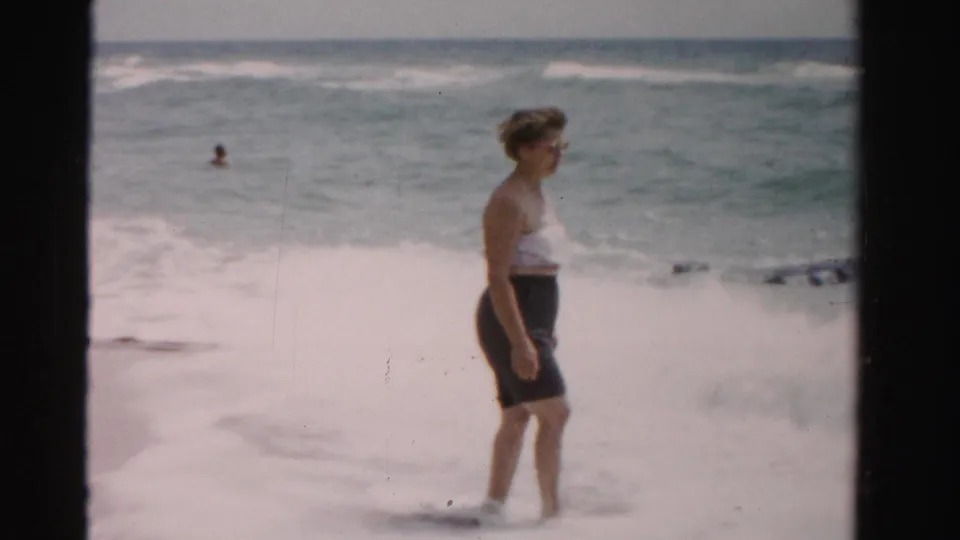 In 1955 Dade County, Florida footage, a woman in beachwear stands at the waterline as waves break behind her.