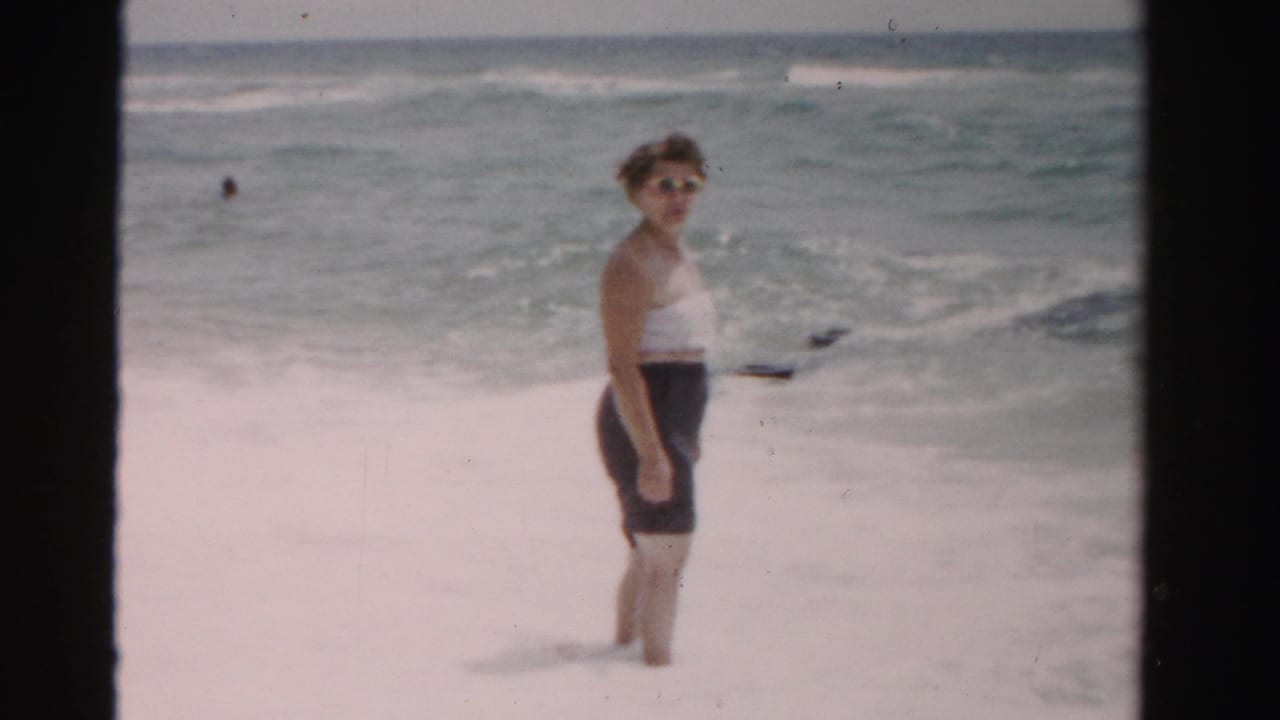 Woman at the shoreline turns partly toward the camera with waves behind her.