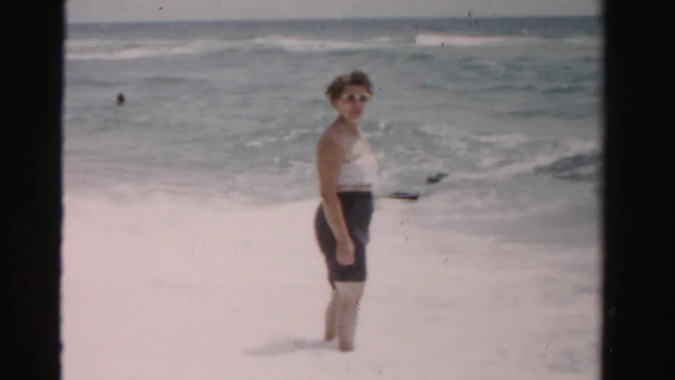 The woman stands in foamy surf in supported 1955 Dade County beach footage, facing partly toward the camera.