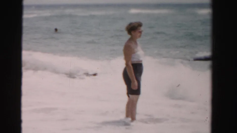 A side view shows the woman standing still in the shallow surf during a 1955 Dade County, Florida beach home movie.