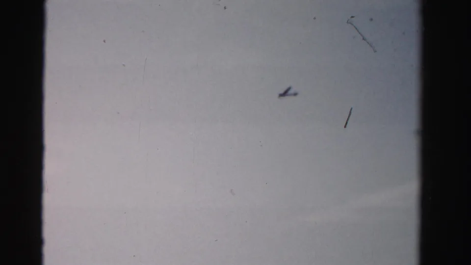 1955 Dade County home-movie frame showing a small airplane silhouette against a pale blue sky with visible 8mm film texture.
