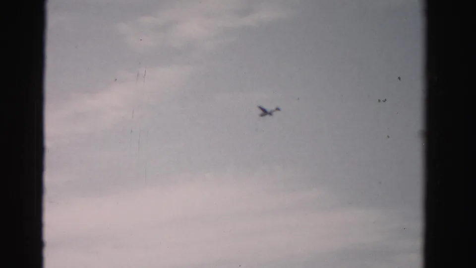 A small airplane silhouette moves across a lightly clouded sky, framed by 8mm gate edges and visible film wear.