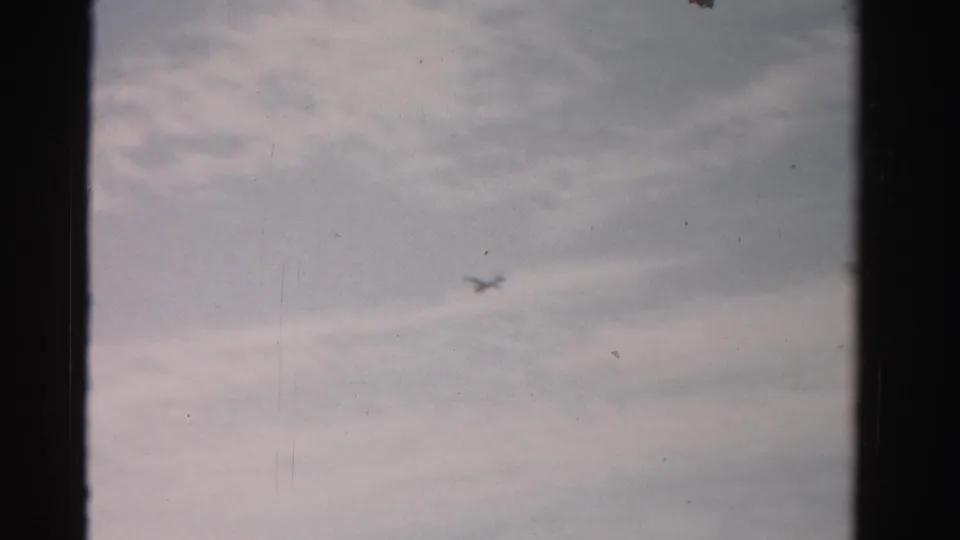 The airplane recedes as a small dark blur beneath thin cloud layers in a 1950s 8mm home-movie frame.