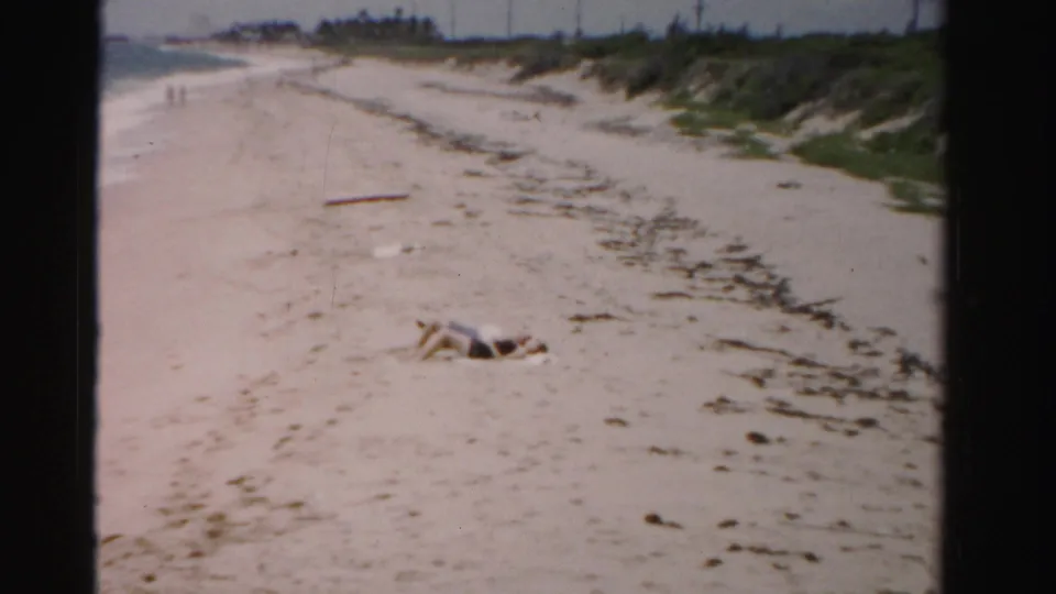 1955 Dade County beach view with a person lying on the sand, seaweed along the shore, dunes, and small figures walking in the distance.