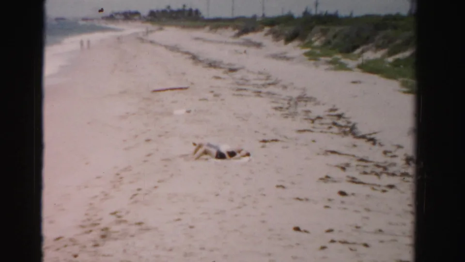 Dade County, Florida beach footage from 1955 showing a reclining person, pale sand, seaweed, low dunes, and distant shoreline walkers.