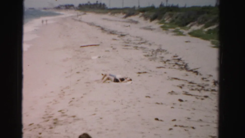 1955 home-movie frame of a Dade County beach with a person lying in the foreground and distant walkers near the surf.