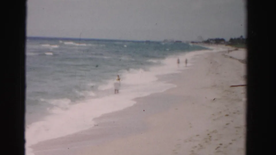 1955 Dade County shoreline view with gentle surf, distant beachgoers, pale sand, seaweed, and dunes receding along the coast.