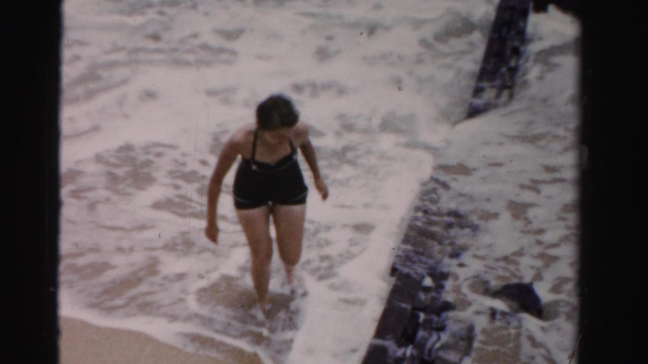 Woman in a black swimsuit walks through shallow surf beside dark pier timbers.