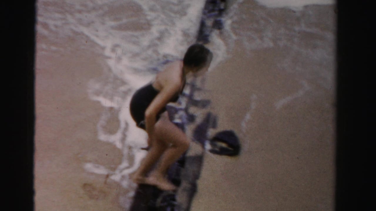 Woman in a black swimsuit climbs over or steps beside a dark pier timber on the beach.