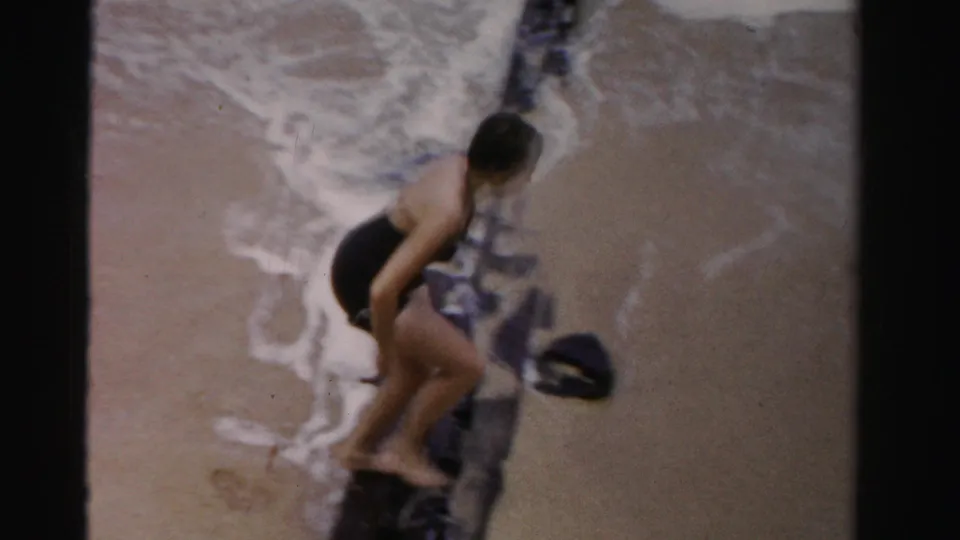 A woman in a black one-piece swimsuit steps near a dark wooden pier support as small waves wash onto the Florida beach.