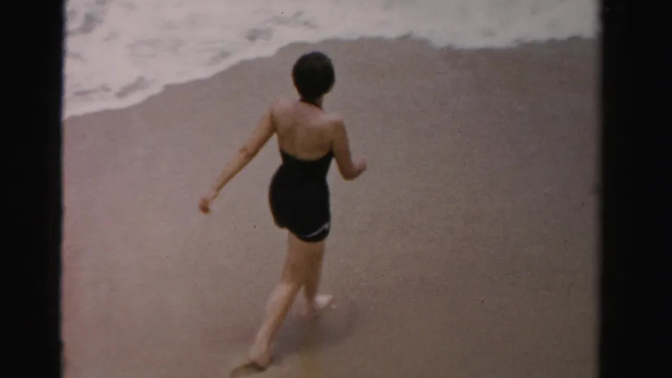 The woman walks along the wet sand away from the waterline, shown from behind in 1950s Florida beach home-movie footage.