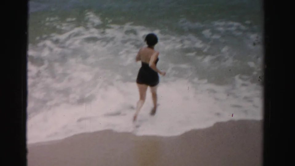 A woman in a black one-piece swimsuit approaches or steps into foamy surf on a Dade County, Florida beach in 1955 home-movie footage.