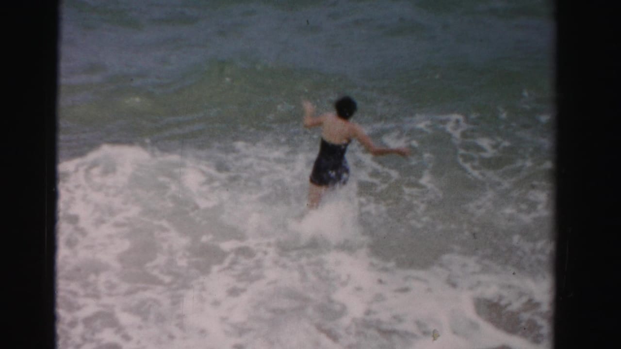 In 1955 Dade County, Florida, a woman in a dark one-piece swimsuit jumps into breaking ocean waves.