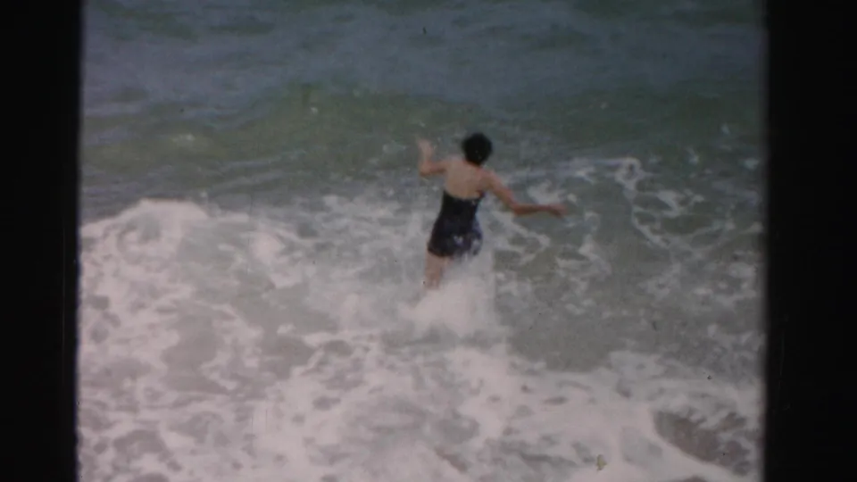 In 1955 Dade County, Florida, a woman in a dark one-piece swimsuit jumps into breaking ocean waves.