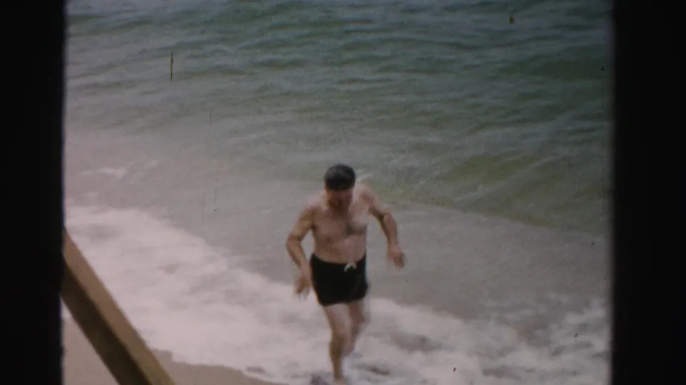 A man in dark swim trunks walks out of shallow ocean water toward the beach, framed by foamy surf.