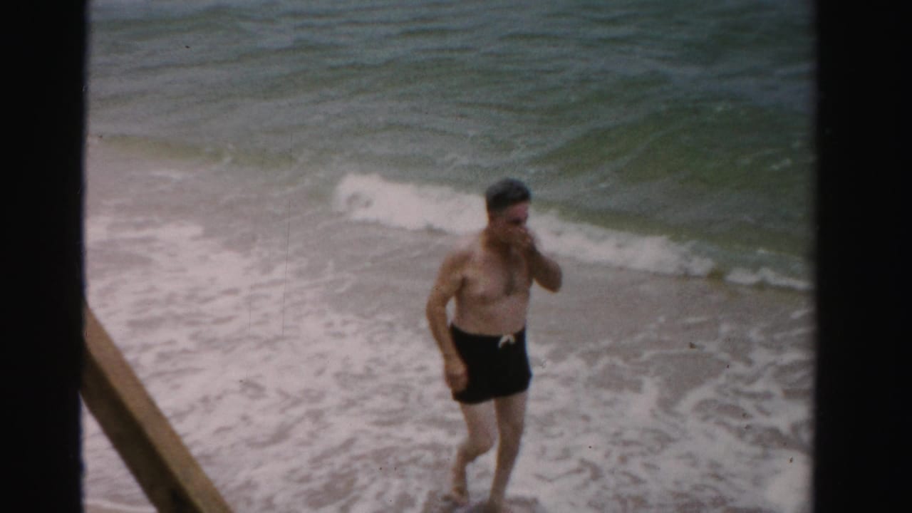 Shirtless man in dark swim trunks wading near foamy ocean waves.
