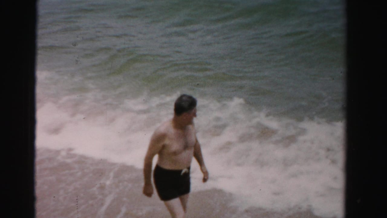 Man in dark swim trunks walking along the waterline with waves behind him.