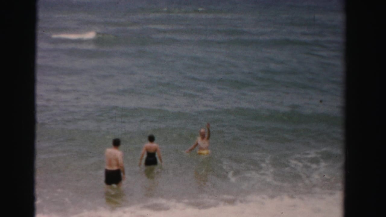 Three adults wade in shallow ocean surf with dark film borders around the frame.