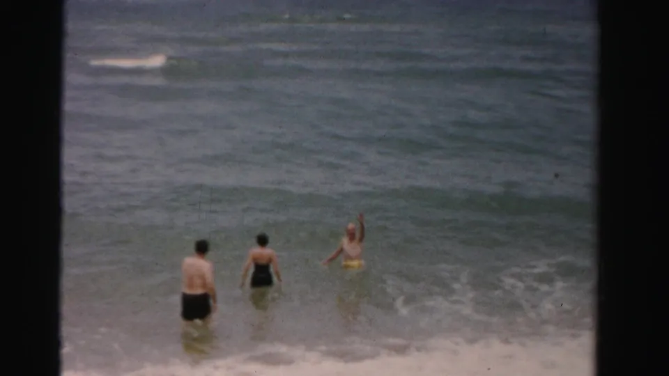Three adults in swimwear wade in the ocean in 1955 Dade County, Florida home-movie footage, with one person raising an arm.