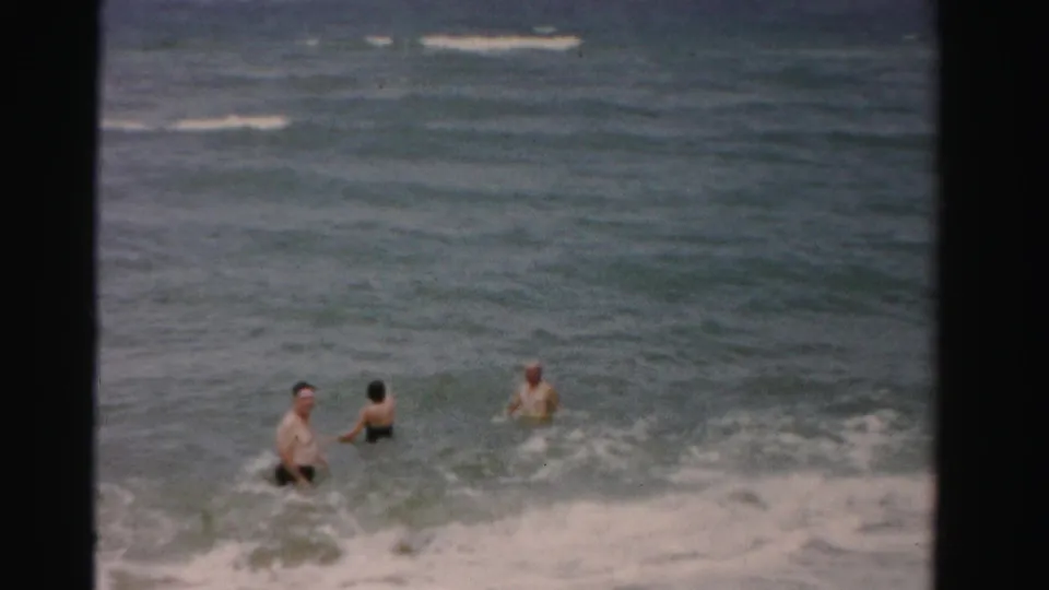 Three adults wade in the ocean off Dade County, Florida, with two figures closer together and another farther right in the surf.